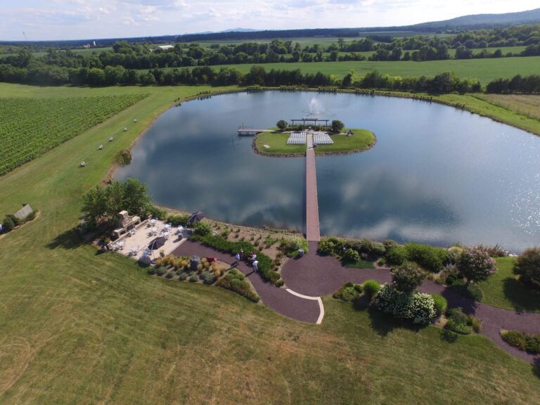 An aerial view of a serene pond with a wooden walkway leading to a small island gazebo. Surrounding the pond are lush green fields and grass. Near the pond's edge is a paved walkway and landscaped garden area with a seating space.