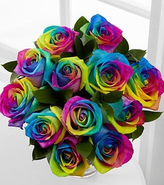 A vibrant bouquet of multicolored roses displayed in a white vase. The petals feature a spectrum of colors, including shades of blue, pink, yellow, purple, and green, creating a striking and playful appearance. The green leaves add a touch of natural contrast.