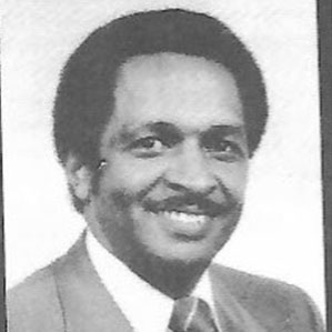 A grayscale photograph of a man wearing a light-colored suit, white dress shirt, and a striped tie. He has short curly hair and is smiling at the camera. The background is plain and white.