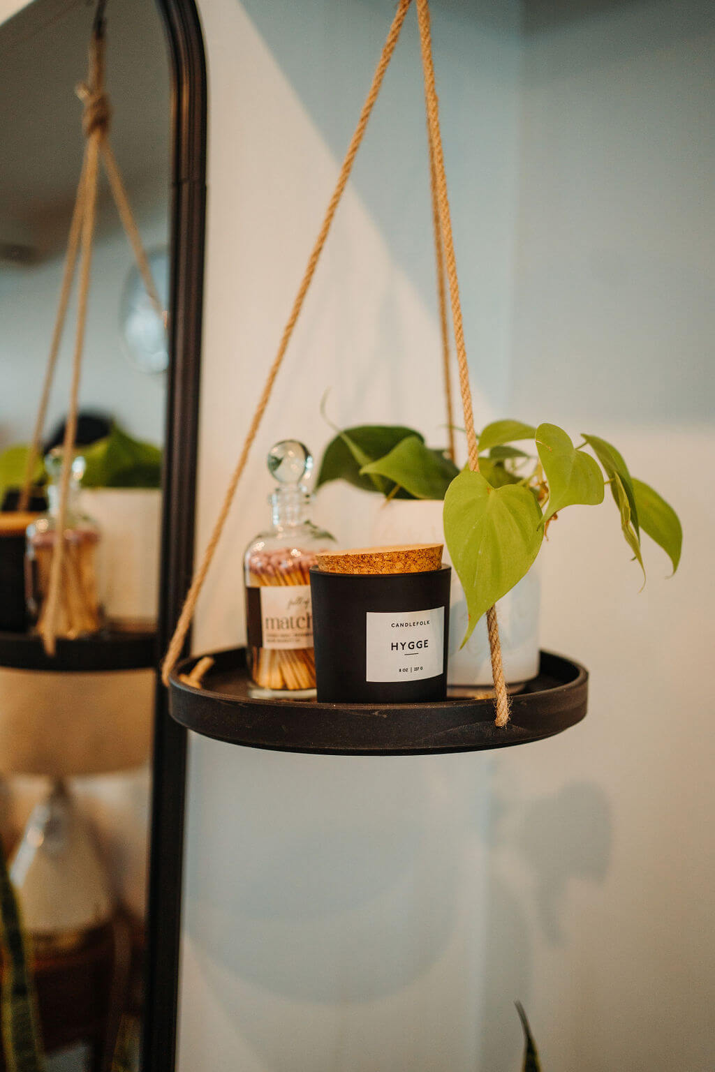 A hanging shelf with rope supports holds a potted plant with green leaves, a glass bottle with a cork stopper, and a small black candle labeled "Hygge." The shelf is positioned next to a mirror and a lamp is partially visible in the background.