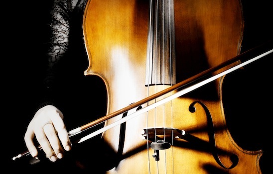 A close-up of a person playing a cello. The image focuses on the torso of the instrument and the hand of the cellist, who is holding the bow and gently pressing the strings. The lighting casts a warm glow on the wooden surface of the cello.