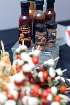 A display of Shawn's barbecue sauces and spices is arranged on a table. In the foreground, there are skewers with cherry tomatoes, mozzarella balls, and herbs. The condiments are neatly organized behind the skewers, creating a colorful and appetizing scene.