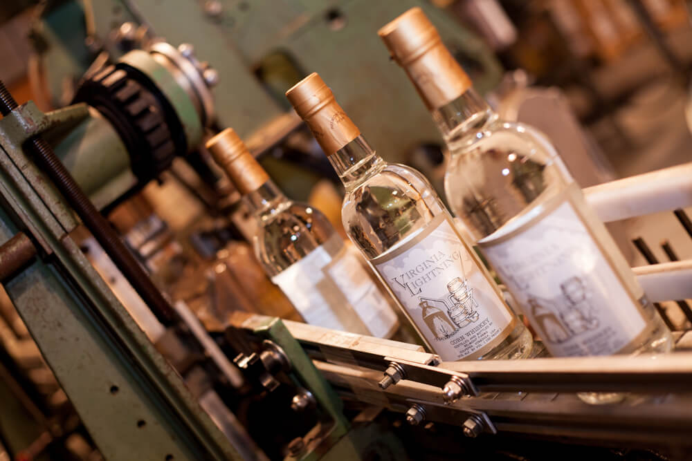 Four bottles of Virginia Lightning corn whiskey are being processed on a distillery production line, moving past metal and mechanical equipment. The bottles are clear with beige caps and labels featuring a corn stalk and the product name.