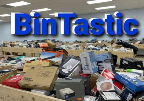 A large room filled with wooden bins, each overflowing with various electronic gadgets, kitchen appliances, and miscellaneous items in boxes. The word 