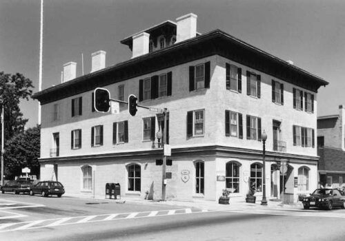 A historic four-story brick building with black window shutters stands at a street corner with a traffic light. The ground floor has large windows and two mailboxes out front. There are a few cars and a streetlamp nearby, and a tree is visible in the background. Image