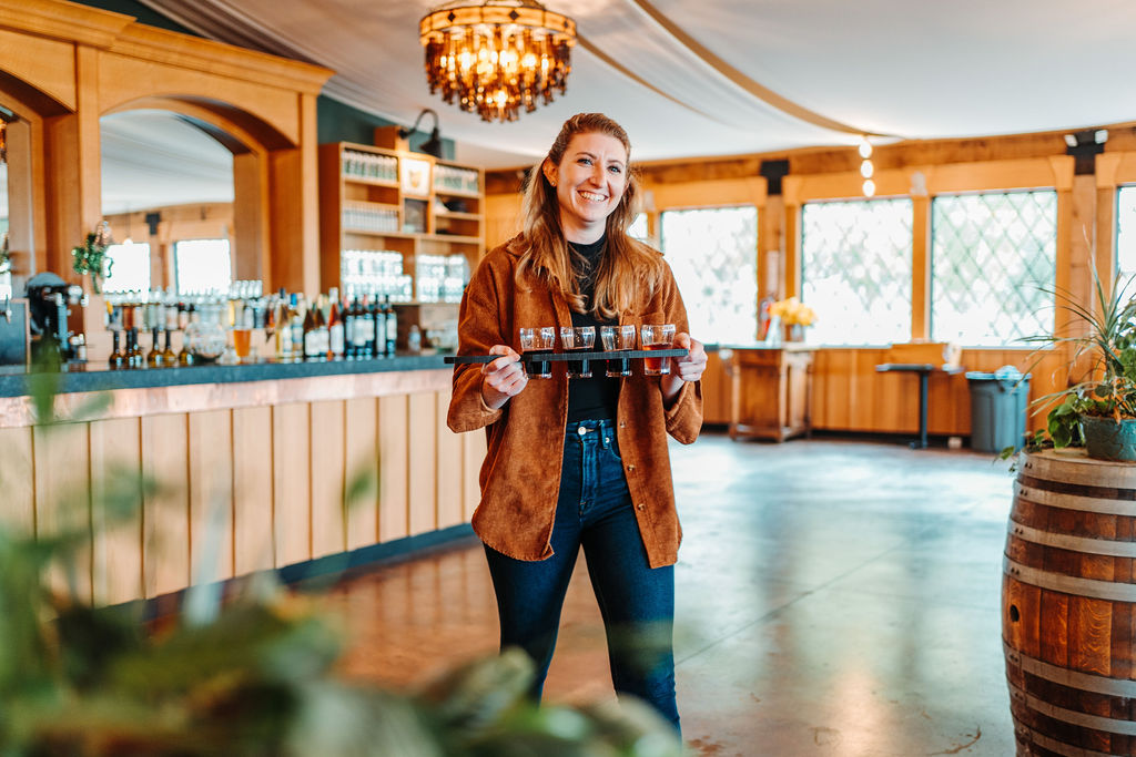 A smiling woman stands in a warmly lit café or restaurant, holding a wooden tray with several glasses. She wears a brown jacket over a black shirt and dark jeans. The interior features wooden decor, a chandelier, and large windows. A wooden barrel is visible in the foreground.