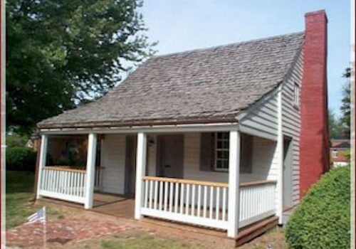 A small, historic wooden house with a steep, shingled roof and a brick chimney on the right side. The house has a front porch with white railing and columns, and a brick pathway leading to the entrance. A large tree is in the background. Image