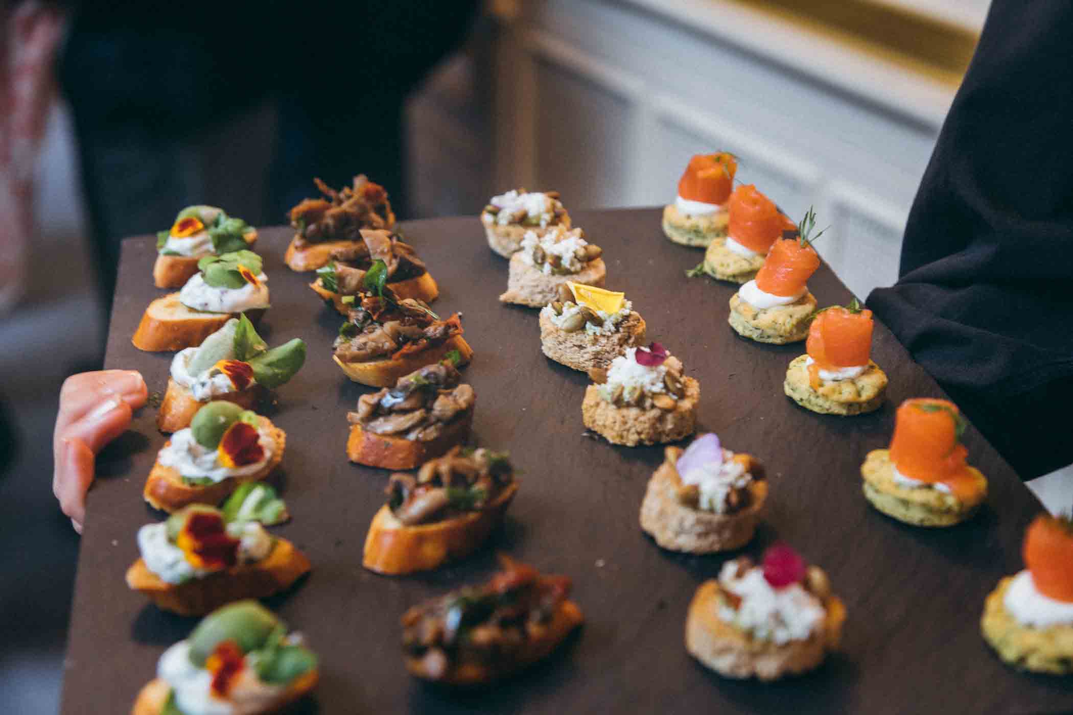 A serving tray filled with various bite-sized appetizers. The food includes assorted bruschetta with toppings like greens, roasted vegetables, and salmon. Each item is garnished with herbs, edible flowers, or small fruit pieces, displayed in neat rows.