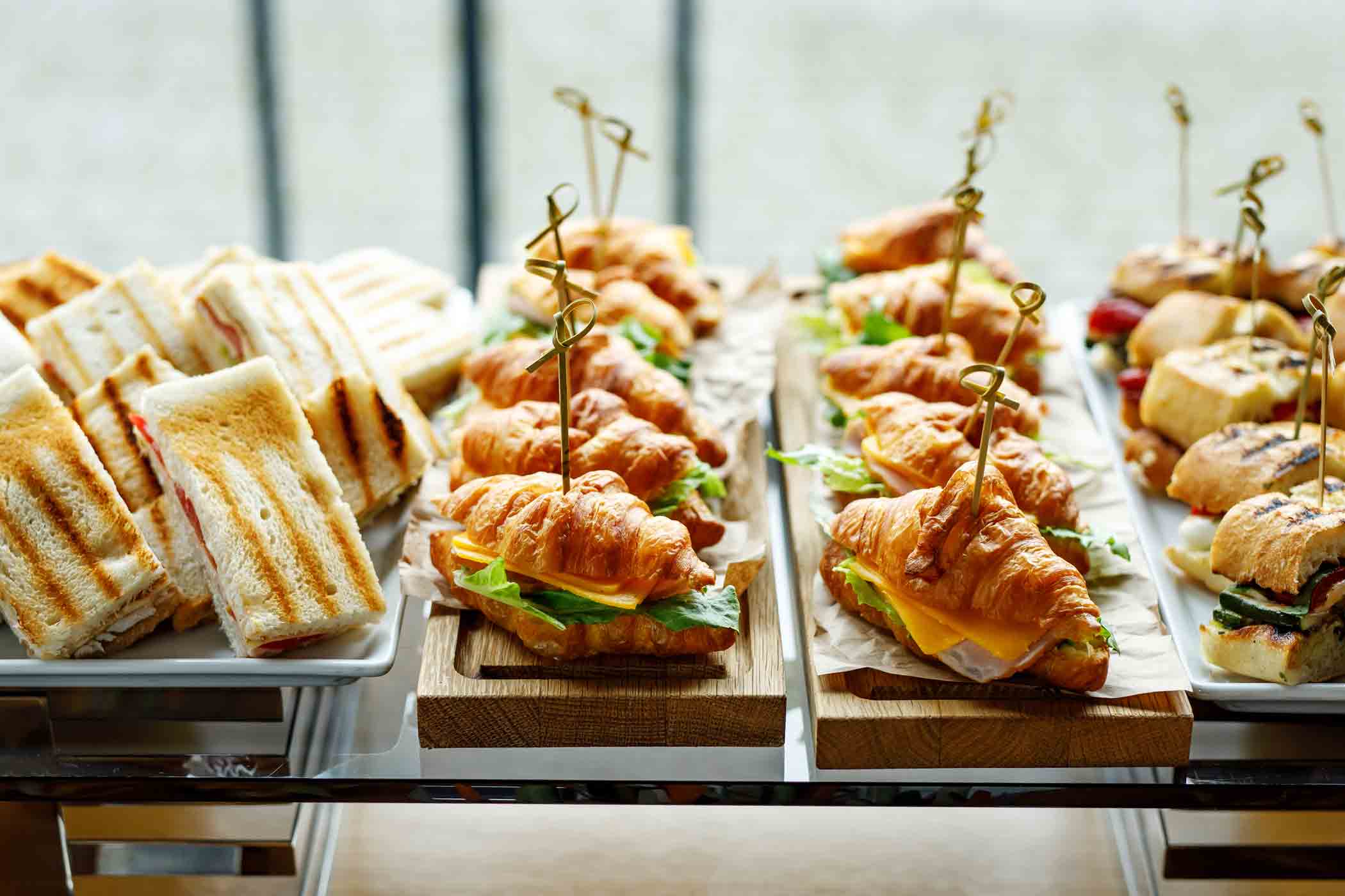 A display of assorted sandwiches includes grilled triangular sandwiches, croissant sandwiches filled with cheese, lettuce, and turkey, and various other sandwich varieties. They are neatly arranged on wooden and ceramic platters on a buffet table.