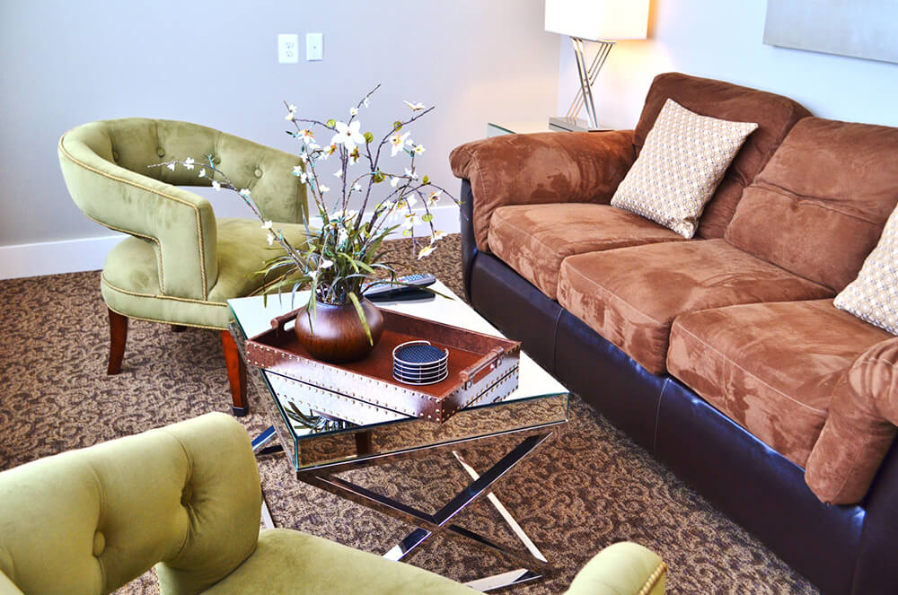 A cozy living room with a brown couch adorned with patterned cushions, two olive green upholstered chairs, a glass coffee table with a potted plant and coasters, and a beige floor lamp. The floor is carpeted in a patterned brown design.