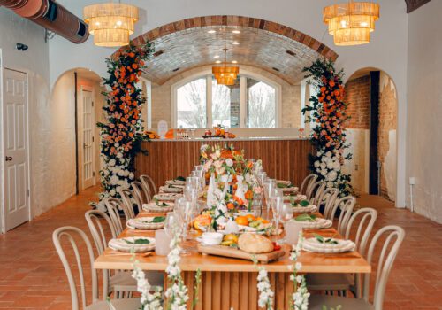 A long dining table set for a meal in an elegantly decorated room with a brick archway. The table is adorned with white flowers, folded napkins, and a variety of dishes. The room features hanging light fixtures and floral arrangements framing the archway. Image