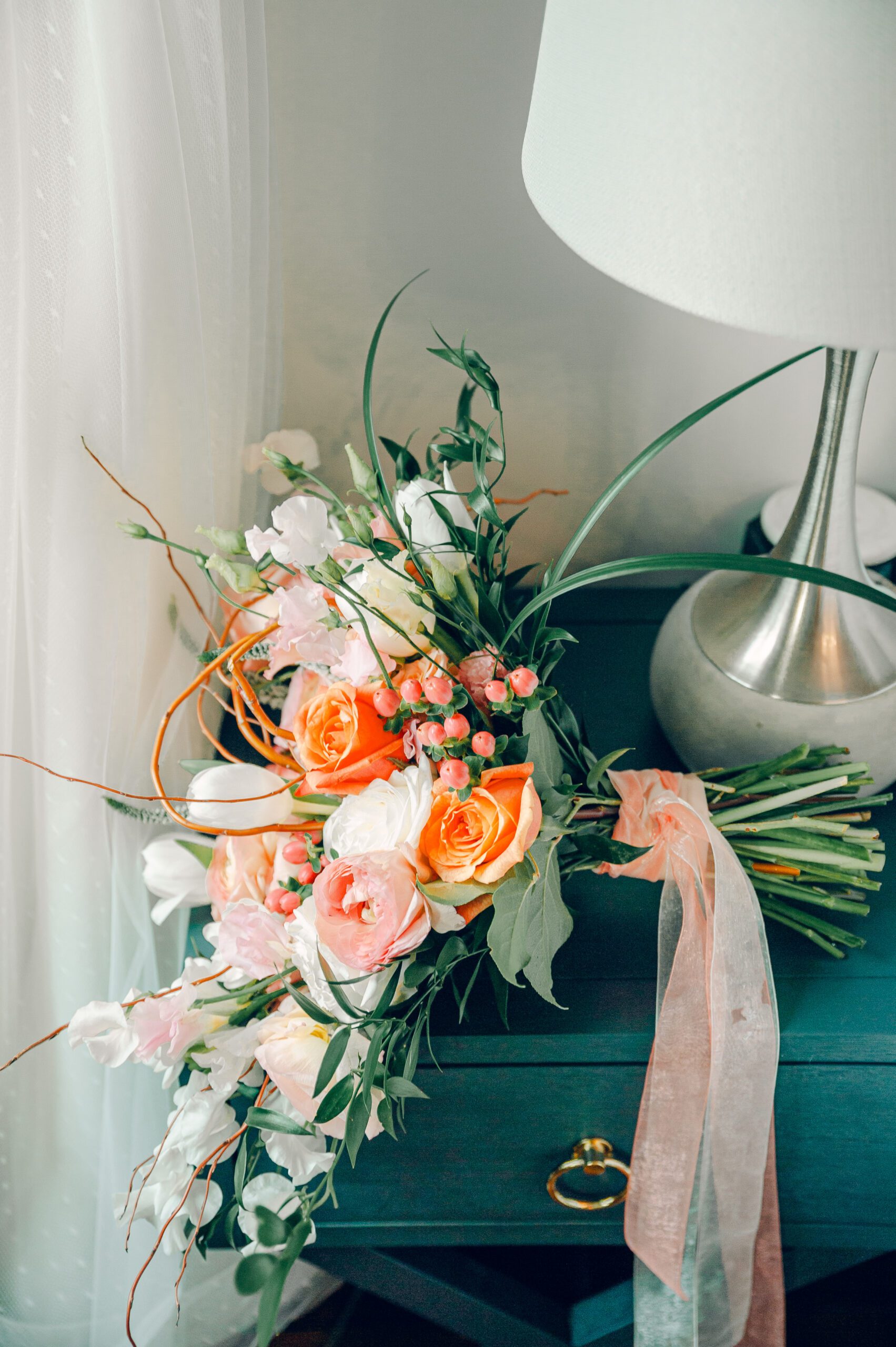 A bouquet of flowers, including pink roses, white lilies, and green foliage, tied with a sheer ribbon, is placed on a dark blue nightstand near a silver lamp with a white shade. White curtains are partially visible in the background.
