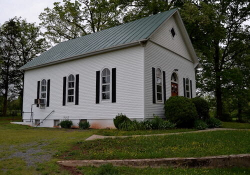 A small white wooden church with a green metal roof and black shutters stands in a green grassy area. The building has arched windows and a single front door. Trees surround the church, and a small pathway leads to the entrance. Image