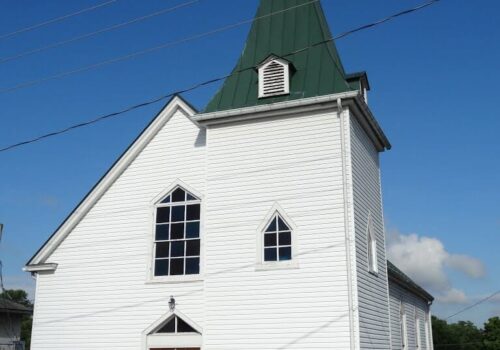 A white church with a green steeple under a clear blue sky. The building features three arched windows near the center, a double door entrance with wreaths, concrete steps, and black railings. A sign on the left reads 