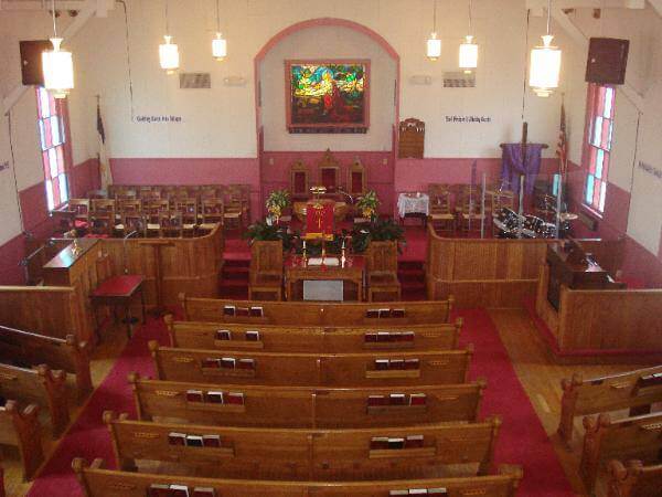 An interior view of a church with wooden pews, a red carpeted aisle, and a pulpit area at the front. A colorful stained-glass window is above the pulpit. Hanging lights illuminate the space, and the walls are adorned with banners and other decorations.