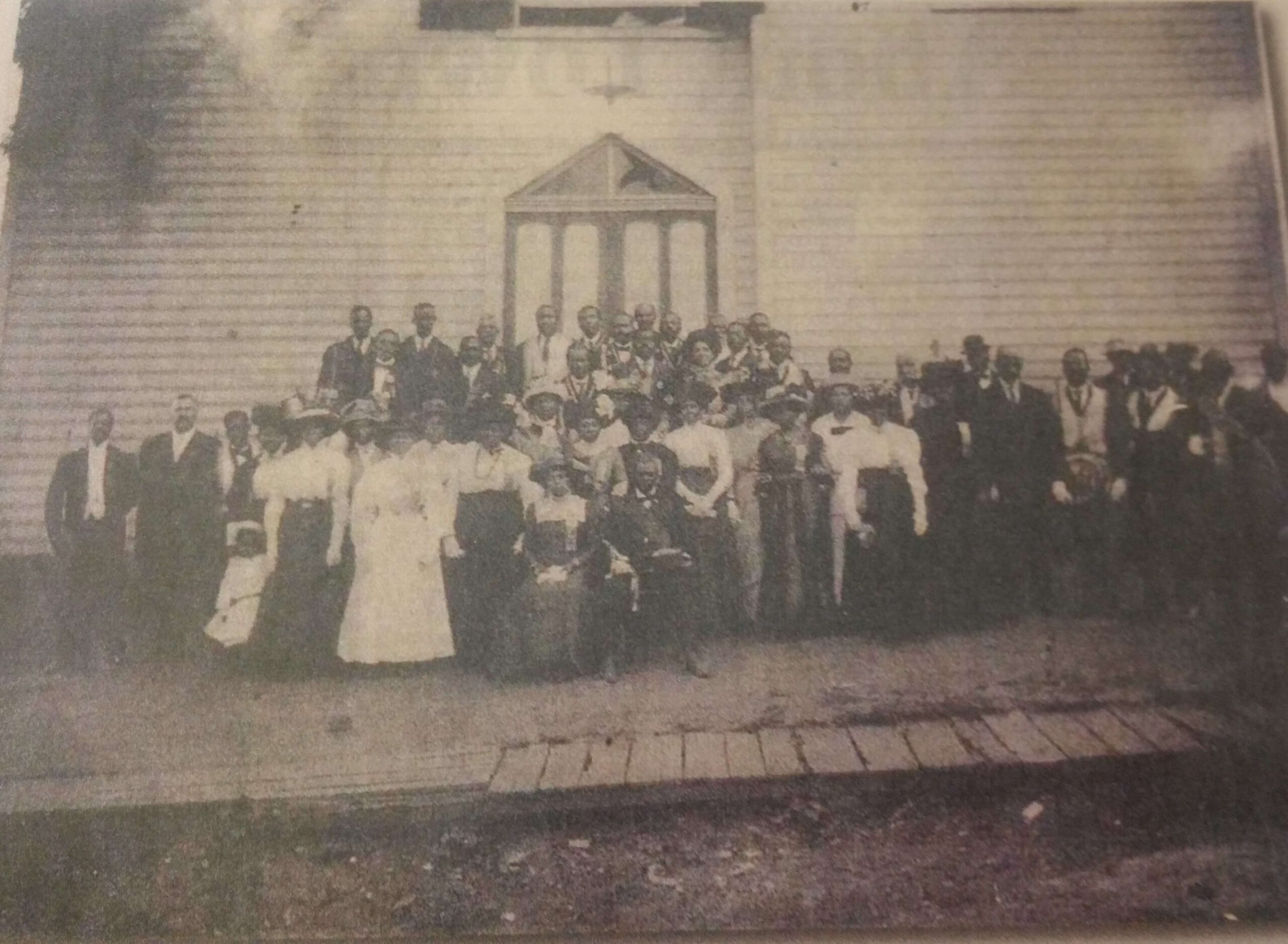 A sepia-toned photograph of a large group of people posing in front of a wooden building with a triangular window above a door. The group consists of men and women, some seated and others standing, dressed in early 20th-century attire.