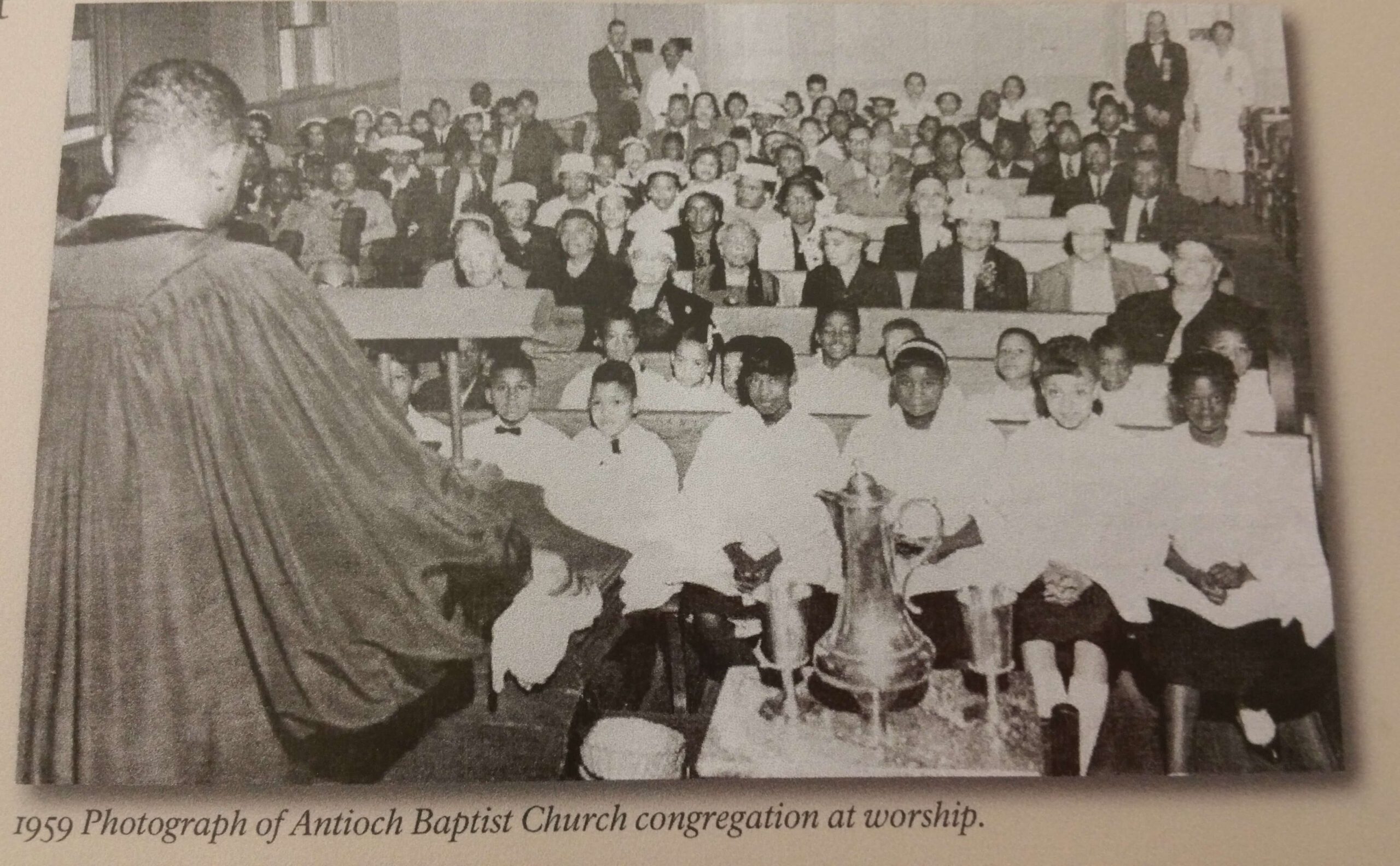 A black and white photograph from 1959 shows a congregation inside the Antioch Baptist Church. The image captures the preacher at the forefront addressing a large group of men, women, and children seated in pews attentively listening.