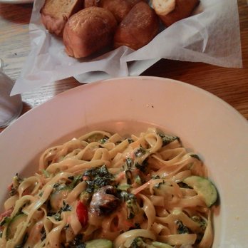 A plate of creamy fettuccine pasta mixed with vegetables, including zucchini and spinach, is served on a wooden table. In the background, there is a basket filled with pieces of garlic bread.