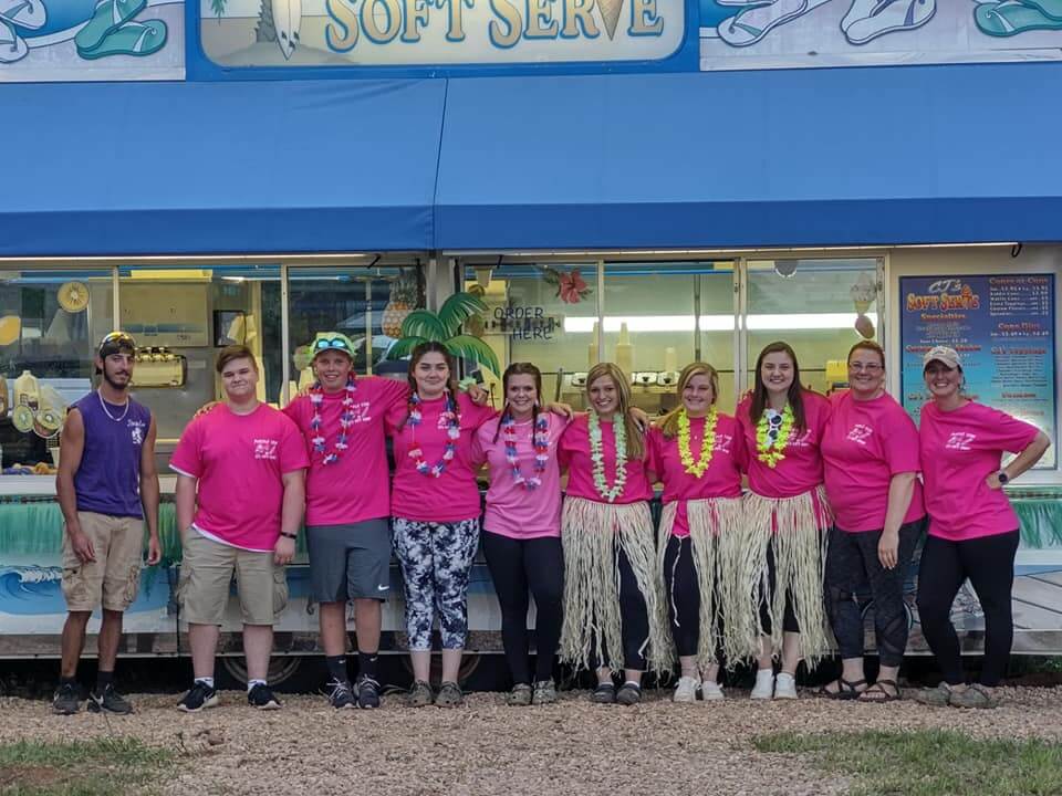 A group of ten people stands in front of a soft serve ice cream stand. They are all wearing bright pink shirts, and some are adorned with leis and grass skirts. The stand has a blue awning with the words 