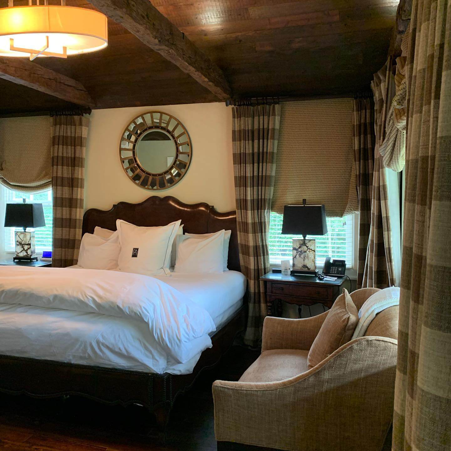 Cozy bedroom with wooden ceiling and beams, a large bed with white linens and pillows, flanked by nightstands with black lamps. A round, decorative mirror hangs above the bed. Curtains cover windows, and an upholstered chair sits in the corner.