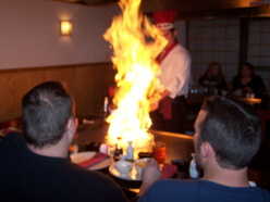 A chef in a red hat and white uniform is performing a teppanyaki cooking show, creating a large flame on the grill. Two people seated at the table are watching the cooking with interest. The setting appears to be a Japanese restaurant.