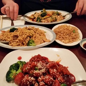 A table covered with various Chinese dishes including a plate of sesame chicken with broccoli in the foreground, a plate of noodles with vegetables and a plate of fried rice in the background. Two hands holding chopsticks and a fork are partially visible.