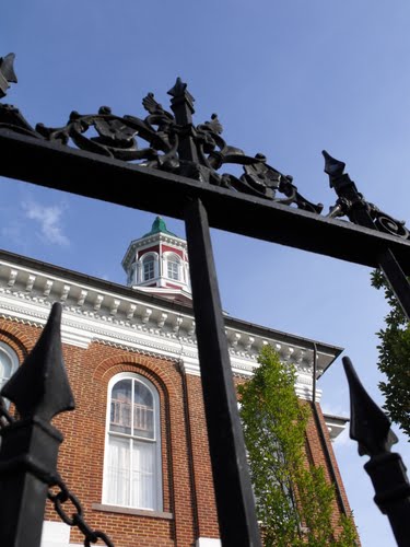 A brick building with arched windows is viewed through an ornate black metal gate. A small cupola with a green, white, and red roof sits atop the building. A tree is visible on the right side under a clear blue sky.