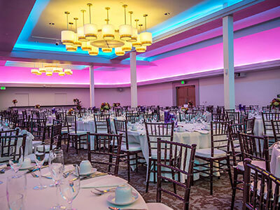 A large, elegantly decorated banquet hall with round tables set for dining. Tables are draped with white tablecloths, set with glassware and centerpieces. The ceiling features modern chandeliers and colorful, ambient lighting in shades of purple and blue.