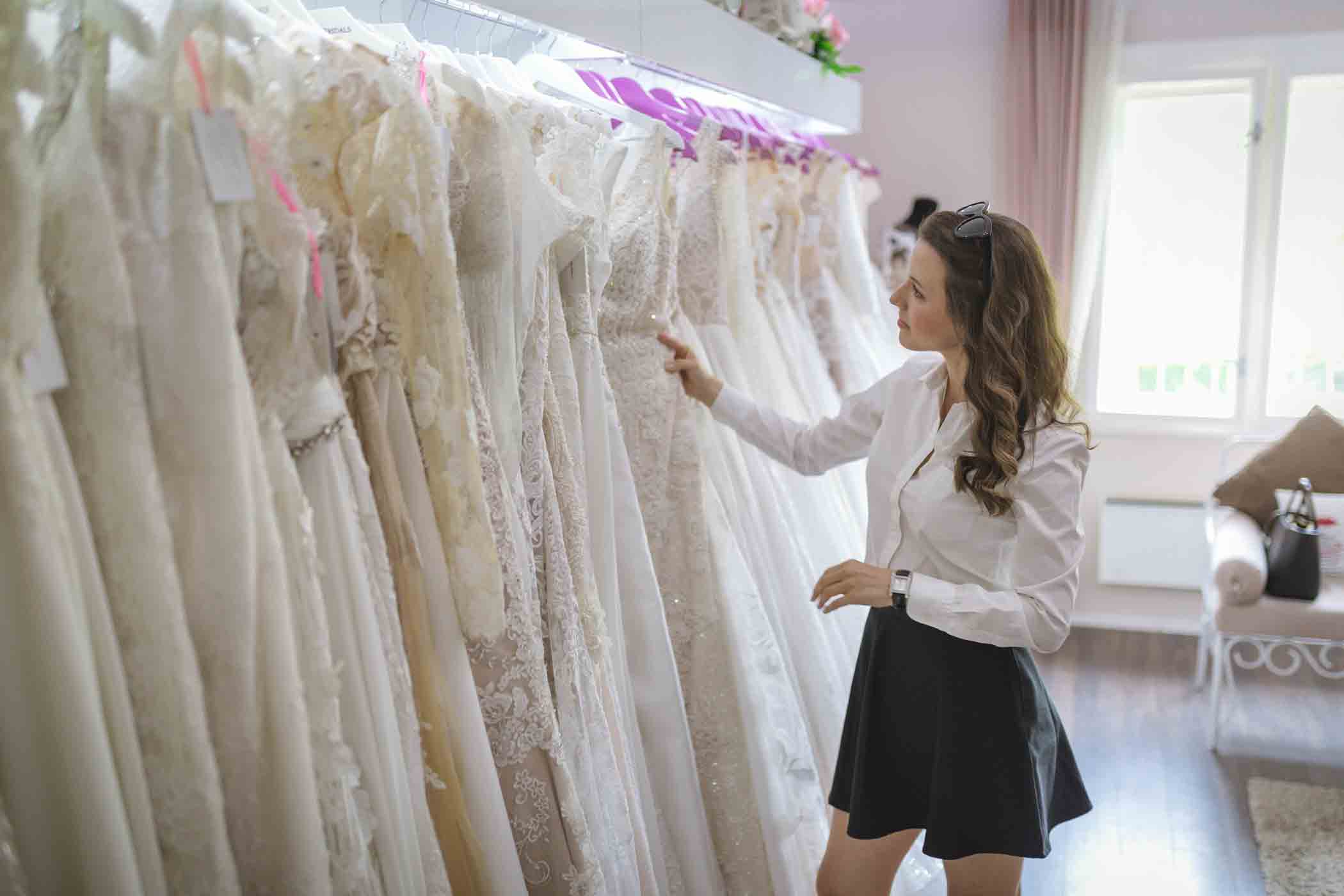 A woman with long hair and wearing a white blouse and black skirt is looking through a rack of wedding dresses in a brightly lit bridal shop. The dresses are white and have intricate lace details. There is a couch and a large window in the background.