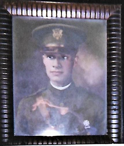 A portrait of a person wearing a military uniform is framed in a dark, ridged wooden frame. The individual sports a peaked cap with an insignia on the front and a high-collared jacket. The photo appears slightly faded and has a reflective glare.