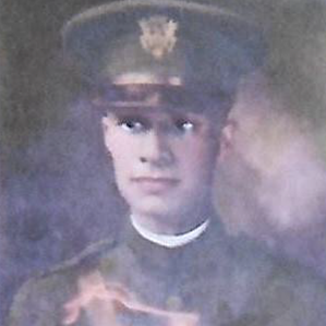 A vintage photograph depicts a young military officer in uniform. He is wearing a peaked cap with an insignia on the front and a high-collared tunic. The image has a faded, sepia tone, giving it an antique feel.