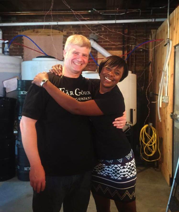 A smiling man and woman stand in a warm embrace. The man is wearing a black t-shirt with white text, and the woman is wearing a black top with a patterned skirt. They are in an indoor setting with industrial equipment and yellow cables visible in the background.