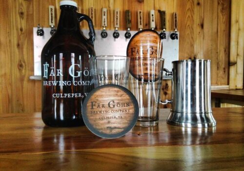 A wooden bar counter displays a growler, two glasses, a metal tankard, and a coaster, all branded with 