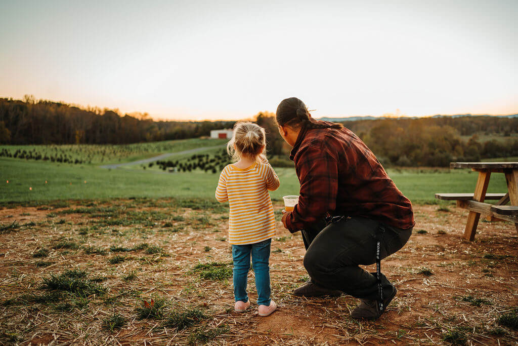 A person wearing a red plaid shirt squats beside a young child in a yellow striped shirt, both looking at the sunset over a vast, green field. The field is dotted with trees and a red barn can be seen in the distance. Nearby, there's a wooden picnic bench.