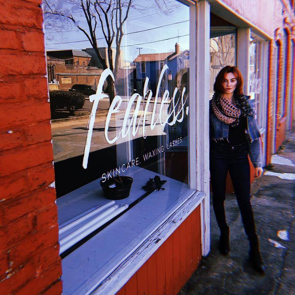 A woman with auburn hair stands outside a storefront with large windows. The window displays the word 