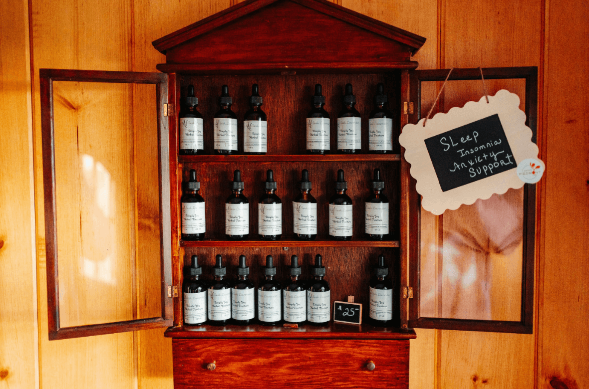 A wooden cabinet with glass doors contains several bottles of herbal tinctures. A small chalkboard sign on the cabinet reads 