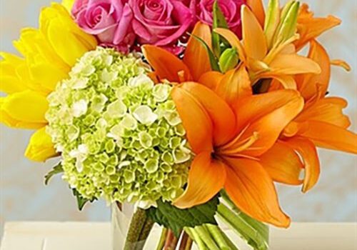 A vibrant bouquet in a clear glass vase featuring yellow tulips, pink roses, orange lilies, and green hydrangeas. The flowers are arranged in a lively mix of colors with light blue and beige background. Image