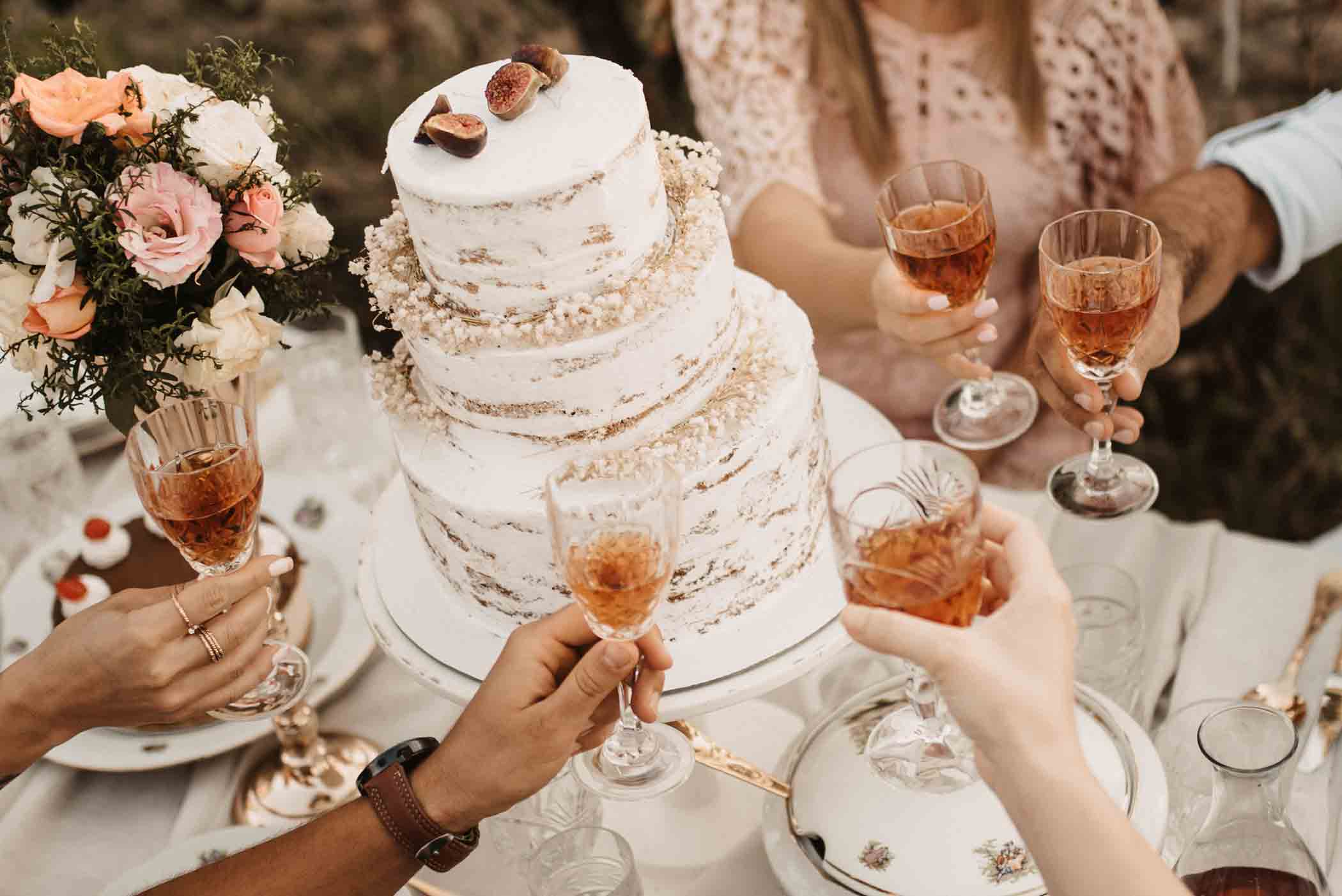 A group of people toasting with glasses of wine around a two-tiered white cake decorated with figs and white flowers. A bouquet of flowers is visible on the left side of the table. The table is elegantly set with plates and cutlery.