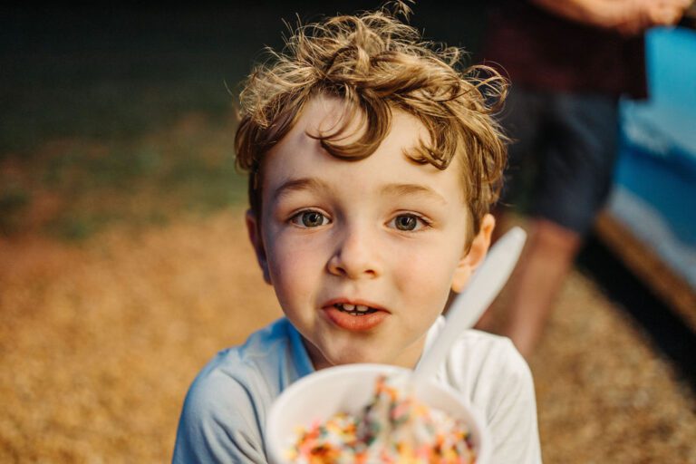 A young boy with curly hair looks at the camera, holding a bowl of ice cream with colorful sprinkles. He is wearing a white shirt and is outdoors, with a blurred figure and background in soft focus. The expression on his face appears curious and delighted.