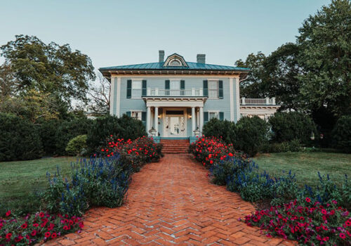 A stately two-story colonial house with blue siding and a green roof, featuring a grand front porch supported by white columns. A vibrant brick walkway flanked by lush gardens of red and blue flowers leads up to the entrance. Green trees surround the property. Image