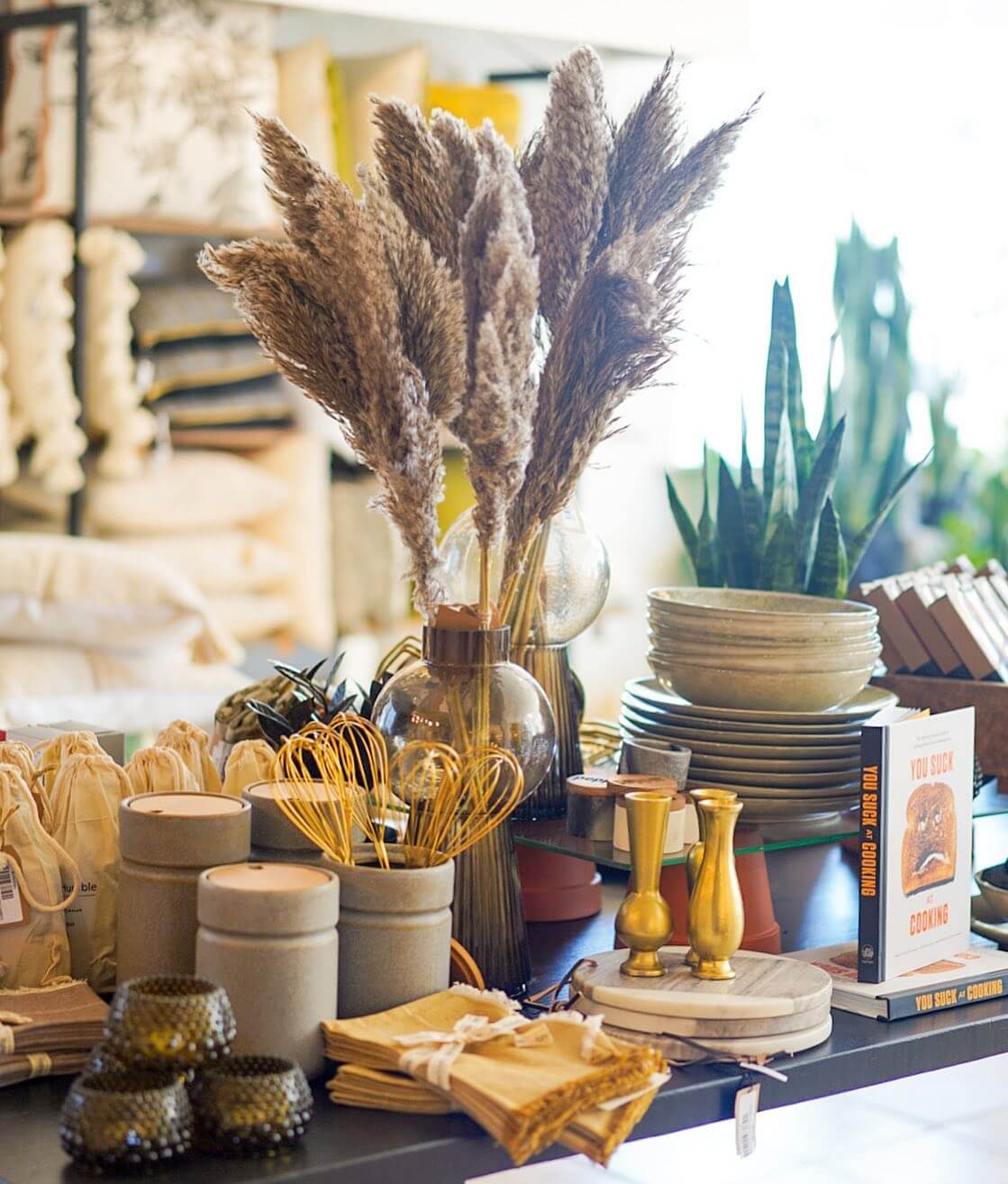 A display table features a variety of stylish home decor items. Prominent items include dried pampas grass in a vase, ceramic bowls, cushions, a cookbook with a cheeky title, brass candlesticks, and potted plants. The overall aesthetic is earthy and contemporary.