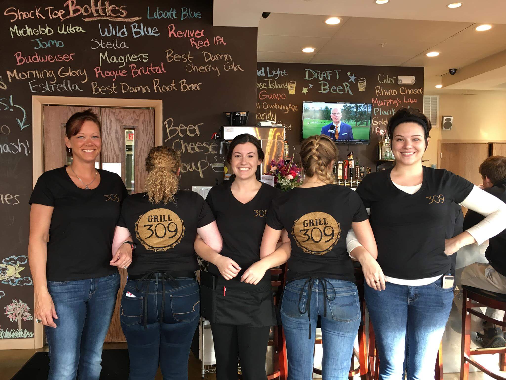 Four women are standing in a restaurant in front of a chalkboard menu. They are wearing black shirts with 