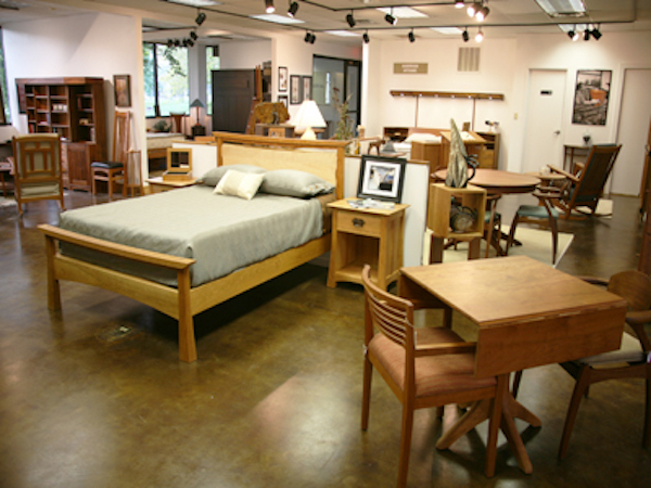 A furniture showroom featuring a wooden bed with gray bedding, a nightstand, a small table with chairs, a dresser, and various other wood furniture pieces. The room has polished concrete floors and is well-lit with spotlights from the ceiling.
