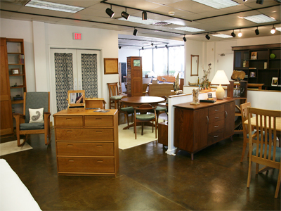 A furniture showroom featuring various wooden furniture pieces, including dressers, dining tables, chairs, and shelves. Warm lighting complements the natural wood tones, and the room has a cozy, organized layout with various accessories placed on the furniture.