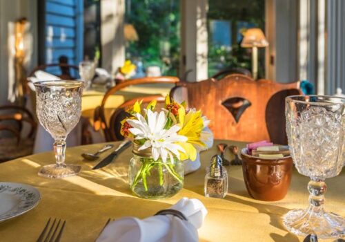 A cozy dining setup with a sunlit table adorned with a small vase of fresh flowers, intricate crystal glasses, a brown ceramic bowl with sugar packets, a salt shaker, and silverware neatly arranged on a yellow tablecloth. Image