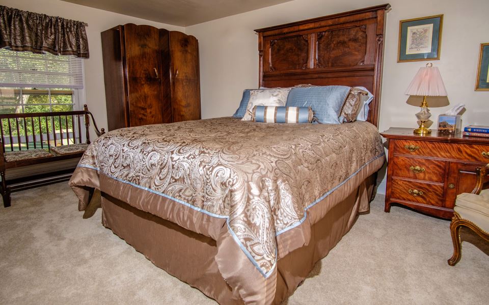 A cozy bedroom featuring a wooden bed with ornate headboard, adorned with gold and blue bedding. A wooden armoire and a bedside table with a lamp, clock, and books are beside the bed. A cushioned bench sits by the window which has sheer curtains. The carpeted floor adds warmth.