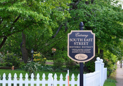 A sign reading "Entering South East Street Historic District" stands by a white picket fence and lush green trees. The plaque mentions the area's historical significance since 1759, urging visitors to respect the private residences and their gardens.
