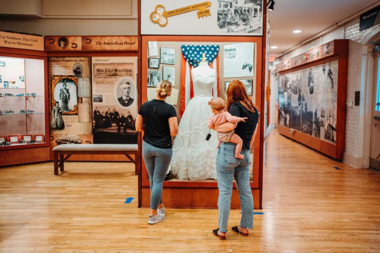 Two women and a baby are looking at a white wedding dress on display in a museum. The exhibit features various historical photographs, text, and artifacts on the wall. The setting is well-lit, with wooden floors and information panels in the background.