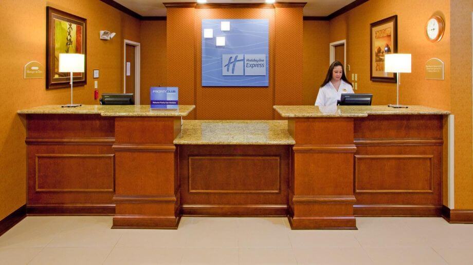 A hotel reception area with a wooden front desk and granite countertops. There are three counter sections, each with a computer monitor. A receptionist is standing behind the rightmost section. The walls are adorned with artwork and a large sign displaying the hotel logo.