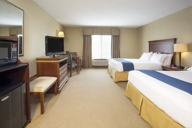 A hotel room with two queen-sized beds, each with white bedding and blue runners. The room includes a flatscreen TV on a wooden dresser, a microwave, a mini fridge, a mirror, and a small table with two chairs by the window. The walls are beige, and the flooring is carpeted.
